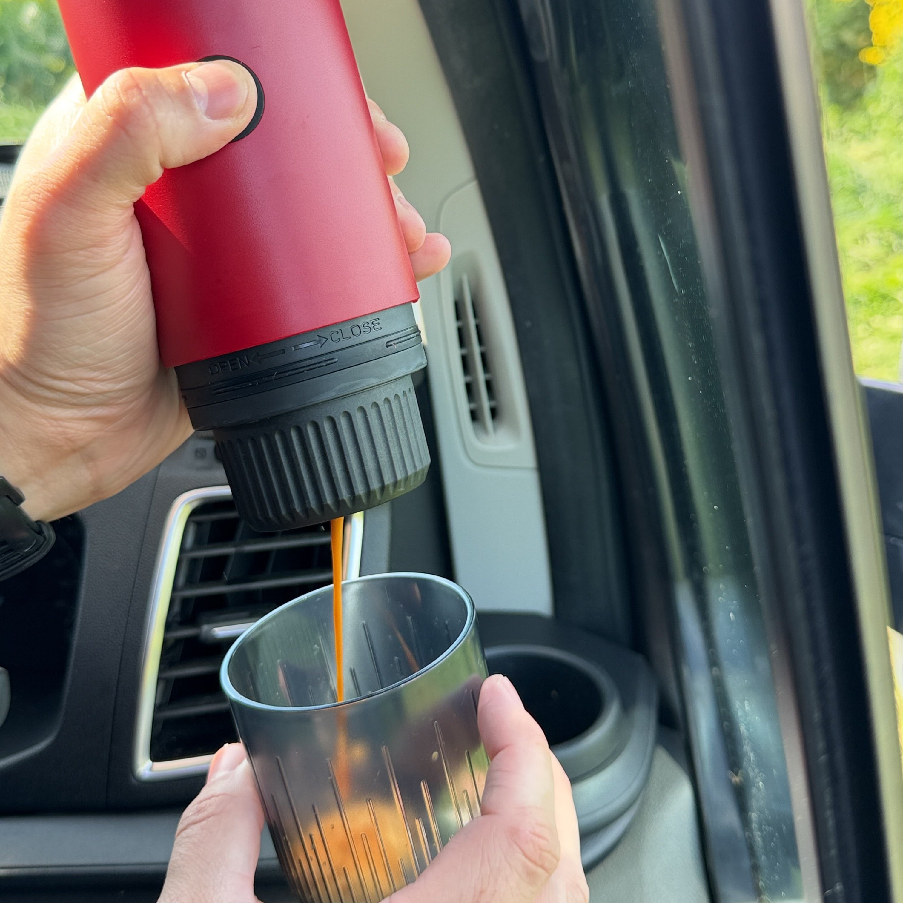 Person pouring coffee from a red and black portable coffee maker into a glass inside a car.
