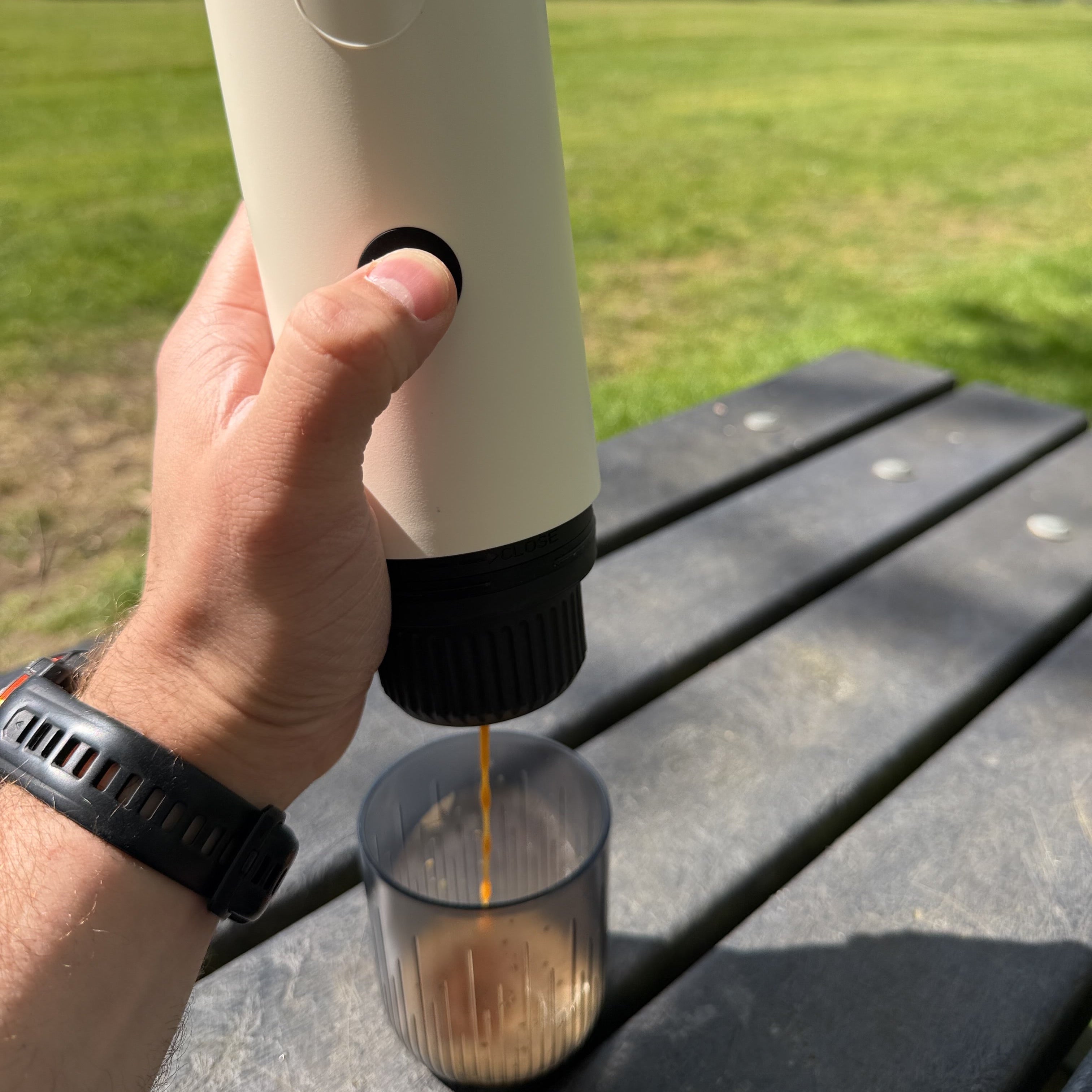 Hand using a white portable espresso maker, extracting espresso showing crema outdoors on a bench